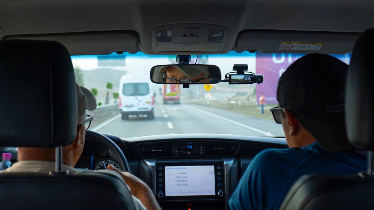 Interior of a car with two people driving on a highway, focusing on dashboard and rear view mirror.